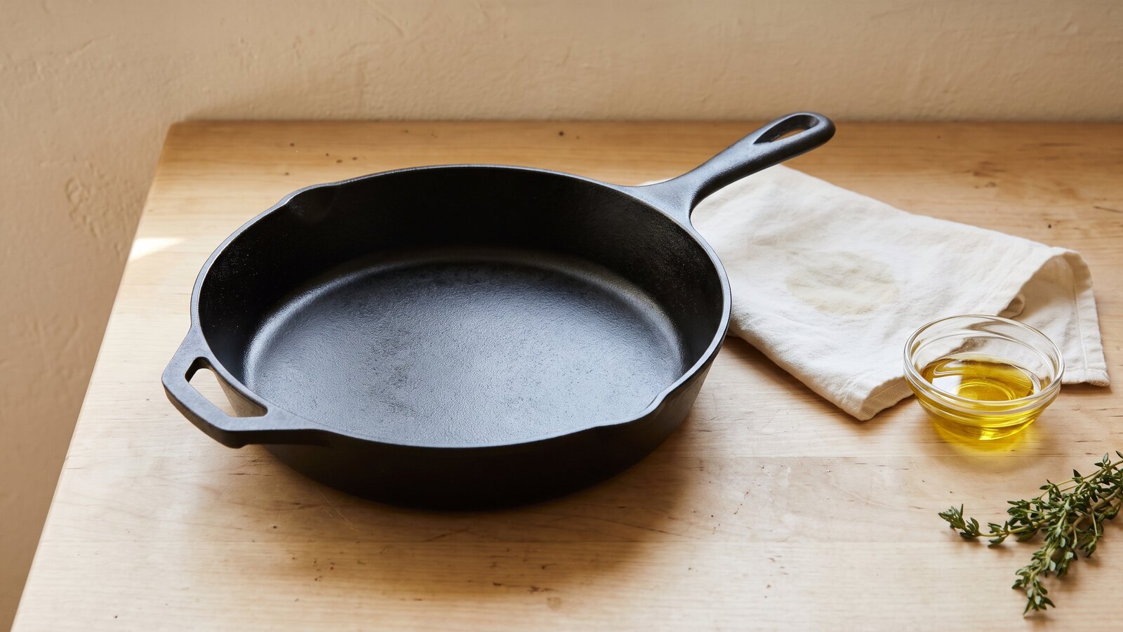 An empty cast-iron skillet with an even, smooth black satin surface on a wooden counter, beside a folded cloth and a small dish of cooking oil.